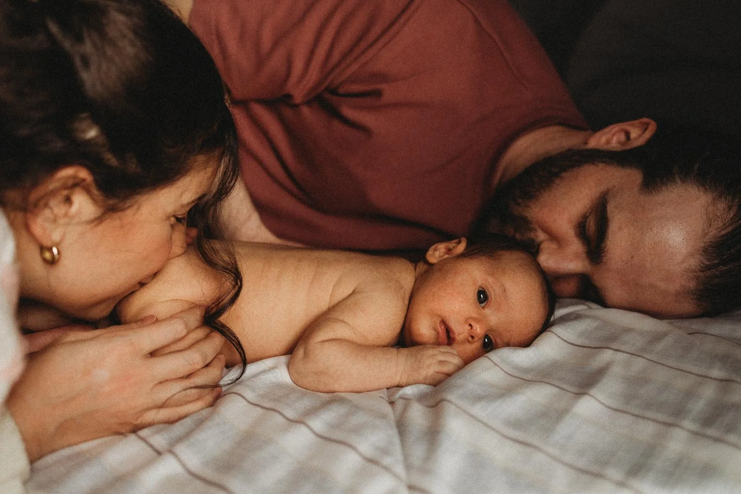 A photo of a mom, a dad and a newborn baby laying down in bed while the mom is gently kissing the baby's butcheek and the dad kissin the baby's head, captured during an in-home newborn photoshoot in Dorval, Montreal