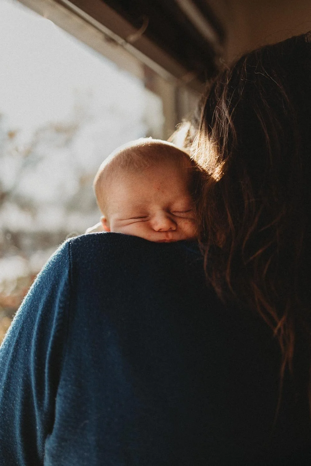 A newborn baby being held by his mom, over the shoulder. The baby's face is a little bit squished and they're in front of a window.