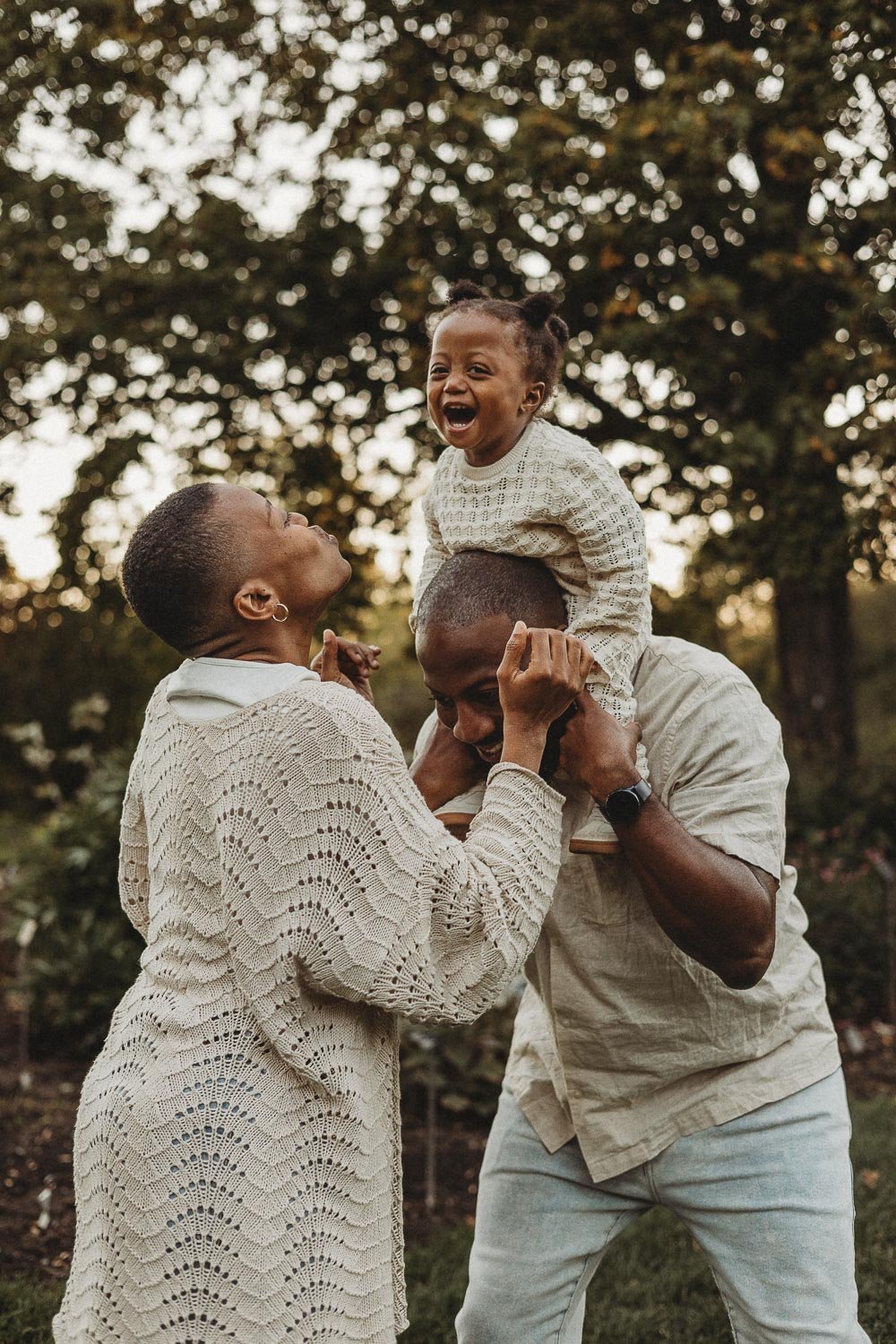 A family of three outdoors, with a woman and man kissing while a young girl is sitting on the man's shoulders, smiling and laughing captured during a family photo session at the Montreal Botanical Garden