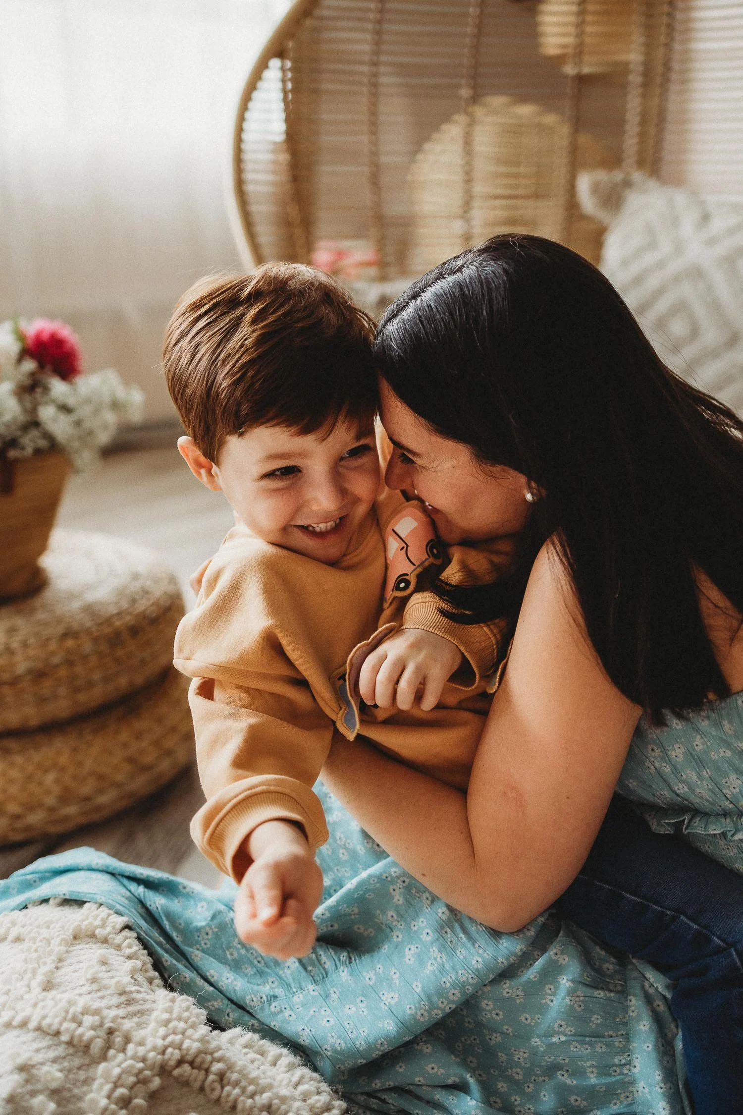 A woman and a young boy giggle and playfully hug, with the woman holding the boy close, both smiling happily, in a cozy room with wicker furniture and a basket of flowers in the background at a studio motherhood photoshoot.