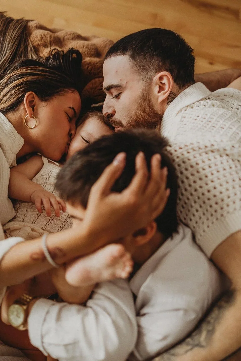A family of mom, dad, a little girl and a boy, laying down in bed while kissing and holding each other during an in-home family photoshoot in Montreal, Quebec