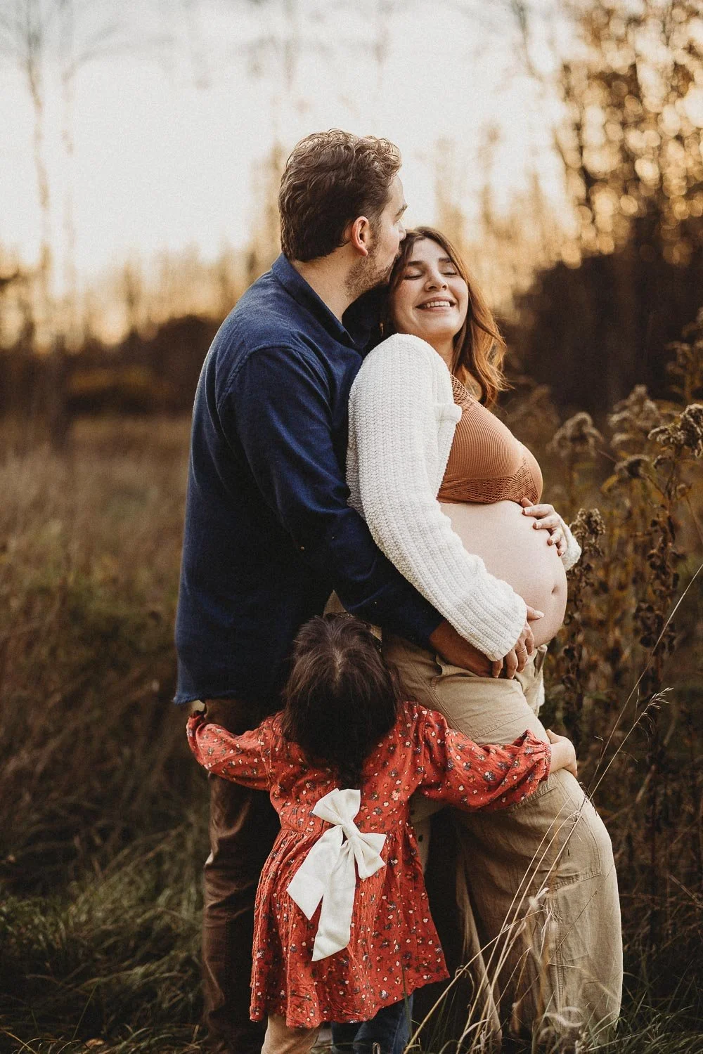 Pregnant woman being held from the back by her partner who is kissing her on the cheek, while both being hugged by their young daughter during an outdoor maternity photoshoot in Boucherville Island