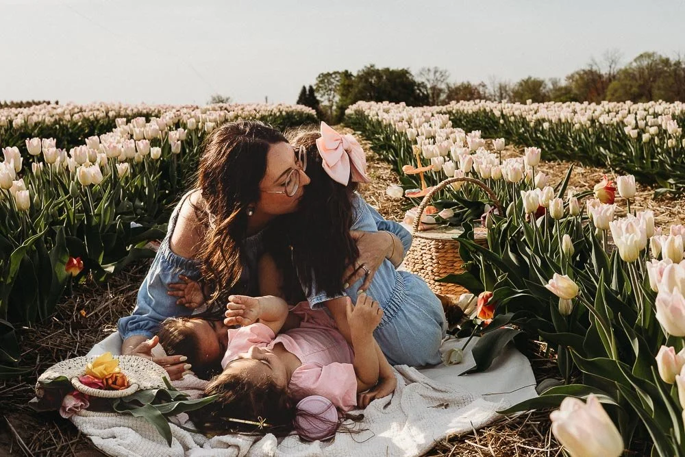 A woman and two young girls in a tulip field during daytime, sharing a hug and intimate moment, with a picnic setup nearby captured during an outdoor motherhood photo session in the Tulip Fields, Laval.
