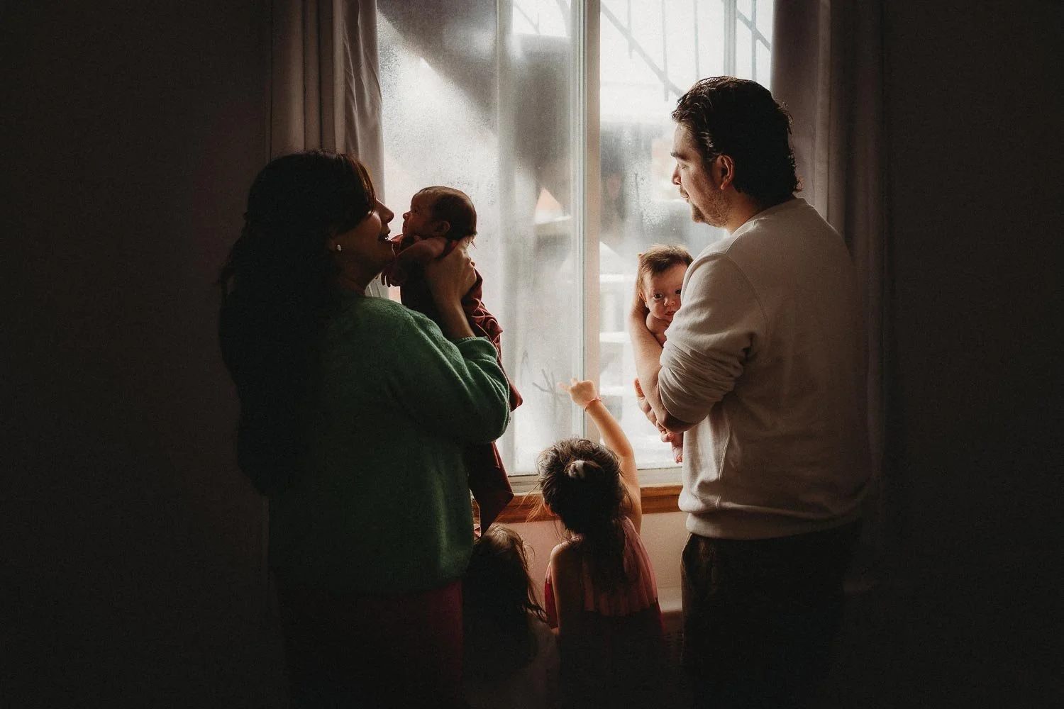 A family of 6 enjoying a quiet moment, parents are each holding a baby while the older daughters are drawing on the window, captured during an in-home newborn photoshoot in La Salle, Montreal