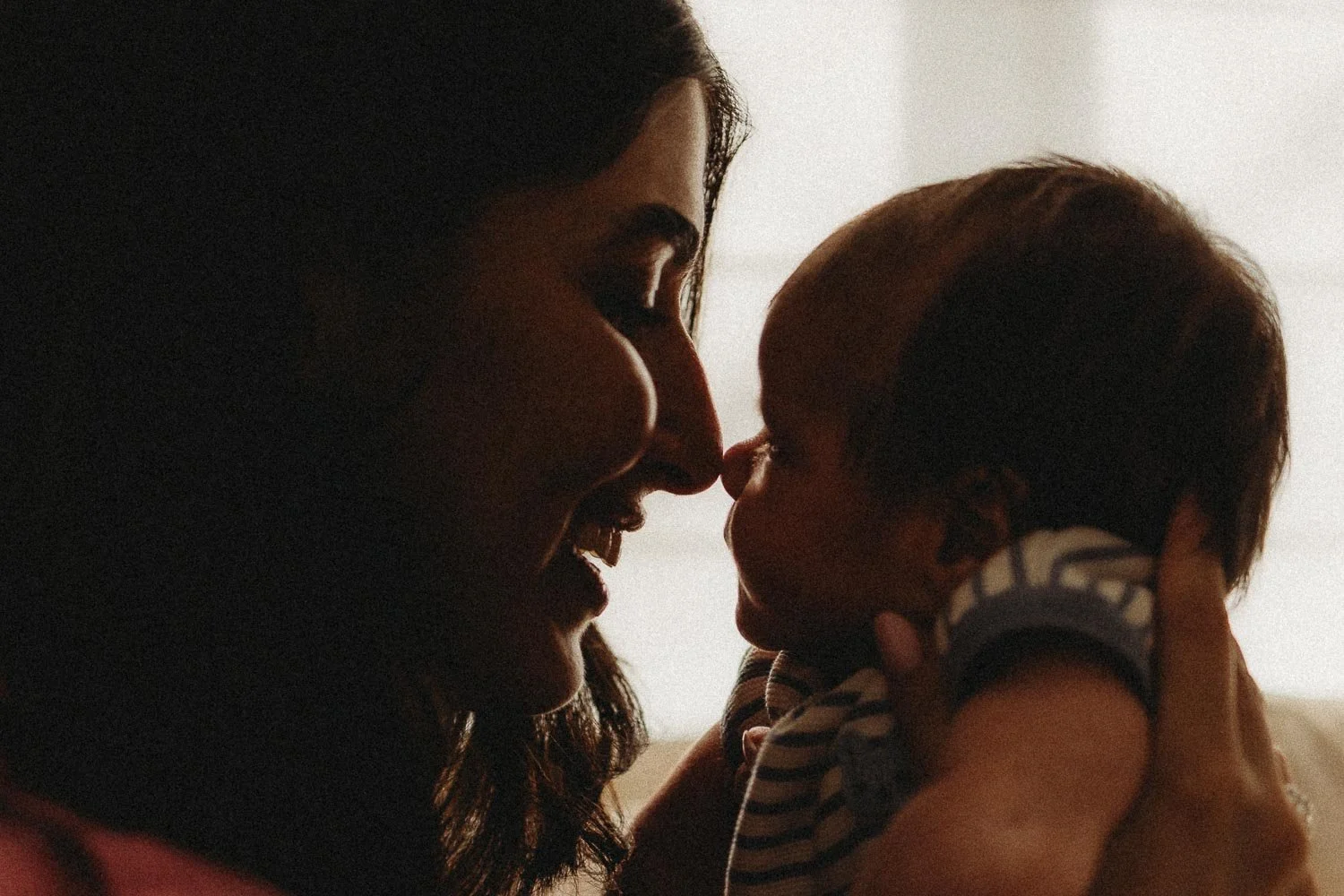 A woman and a baby are close together, touching noses and sharing a tender moment indoors.
