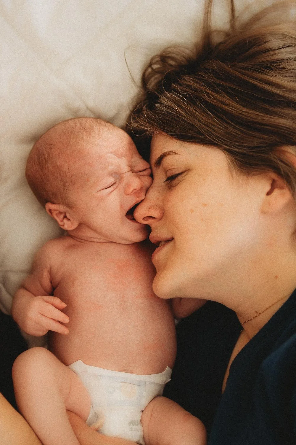 While an in-home newborn photo session, a mom and her baby boy are laying down next to each other, the baby is crying and wanting to suck on mom's nose