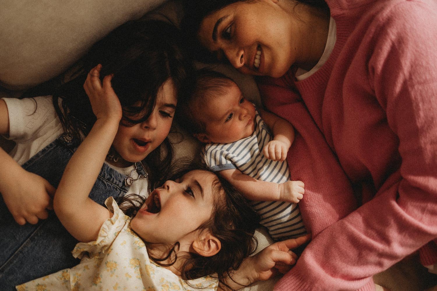A woman lying on a couch with four children, all smiling and looking at each other, enjoying a playful moment during an in-home newborn photoshoot in Hampstead, Montreal