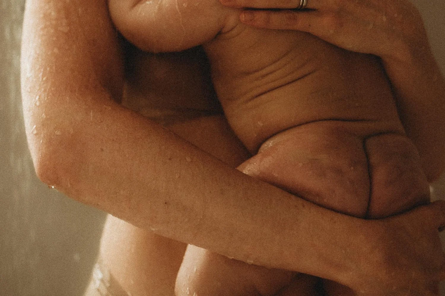 A closed up photo of a mom holding a baby, we see the baby's bottoms and the arms and hands of the mom gently holding him, while taking a shower, captured during an in-home motherhood photo session in Dorval, Quebec