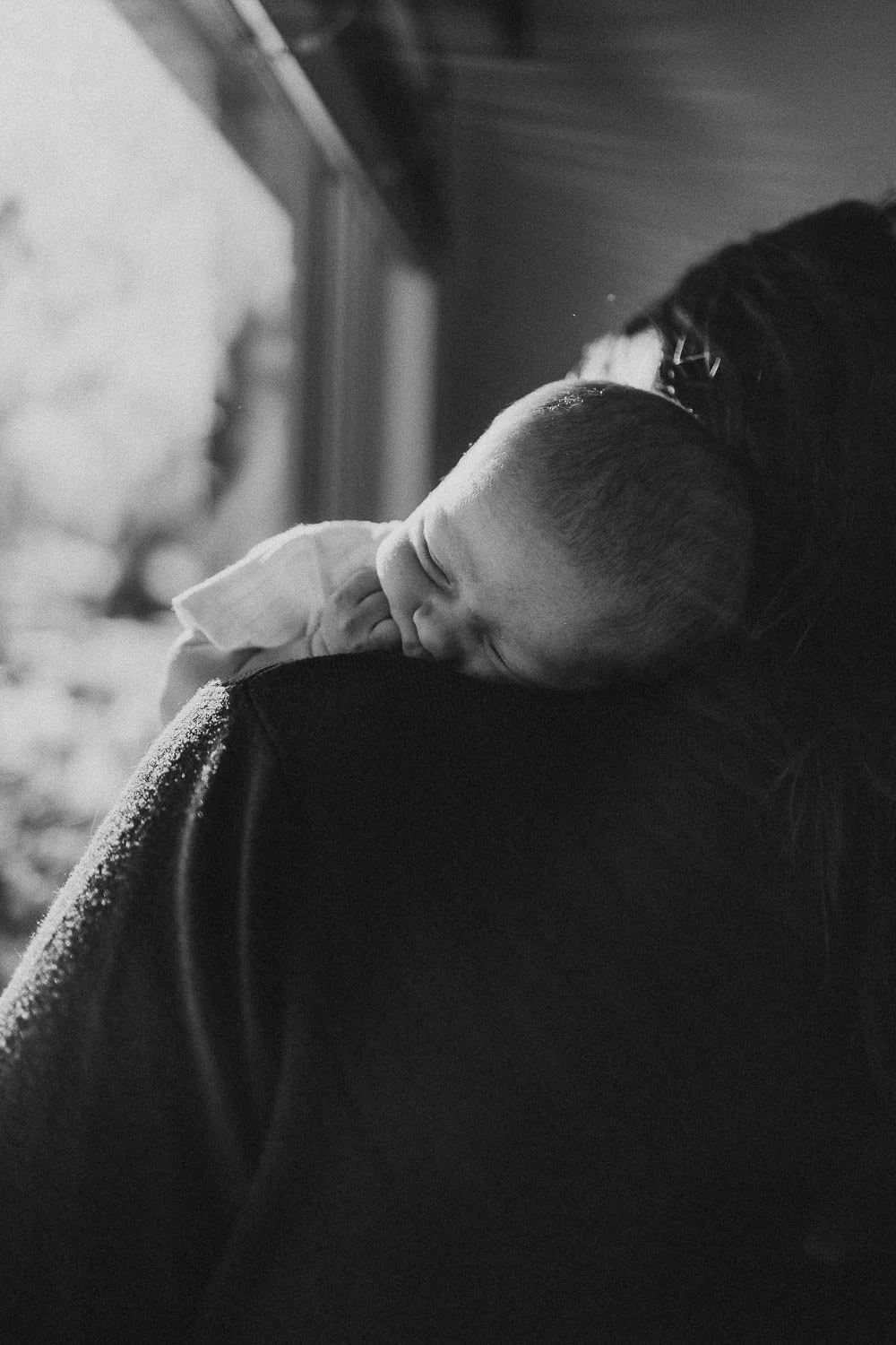Baby with closed eyes and hand in mouth being held by her mom on her shoulder next to a window, captured during an in-home newborn session in Montreal, Quebec