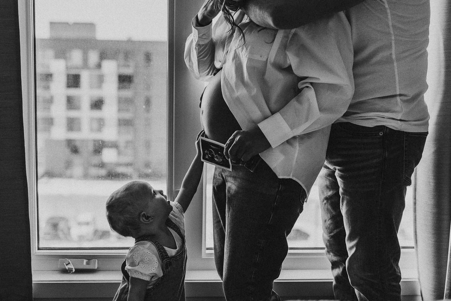 toddler touching her mom's pregnant belly who is holding an ultrasound and being hugged by partner, captured during an in-home maternity photoshoot in West Island, Montreal