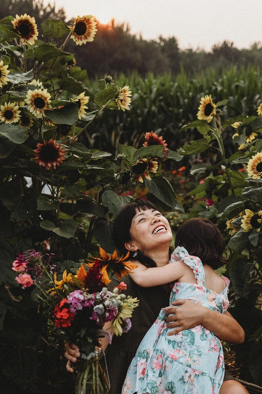 A n asian woman with short black hair is smiling happily while hugging her daughter in a floral dress, surrounded by sunflower plants and colorful flowers during sunset family photo session in montreal.