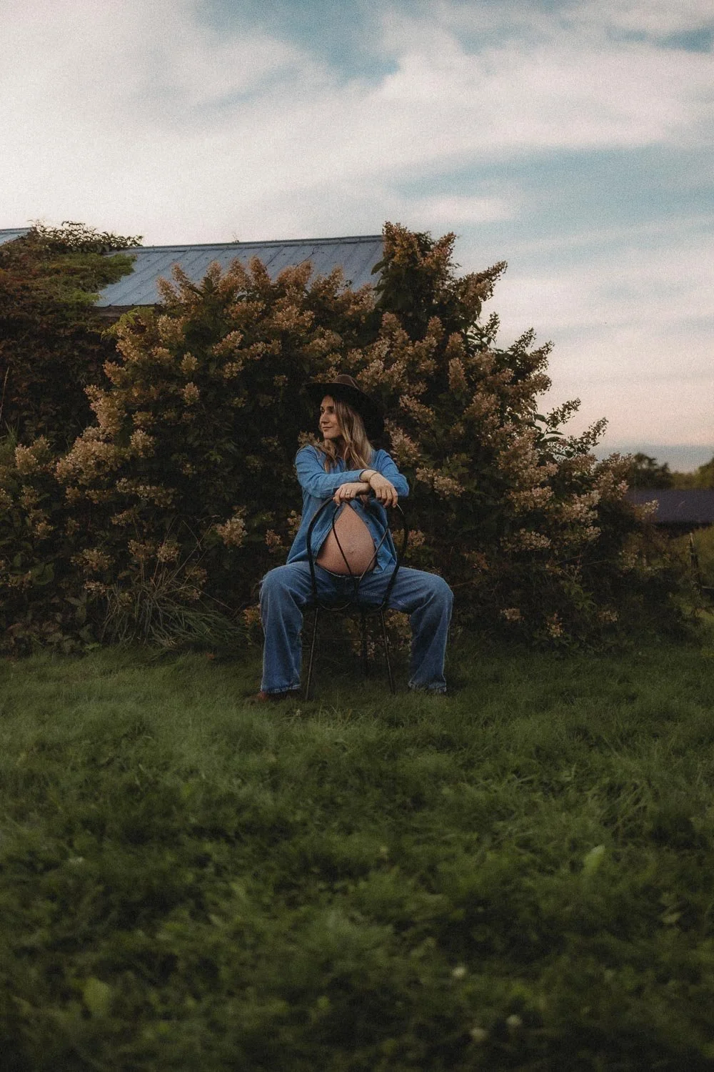 Expectant mother resting both hands on her bump, sitting in long grass beside a flowering shrub captured at golden hour at Le Verger Du Flanc Nord in Quebec