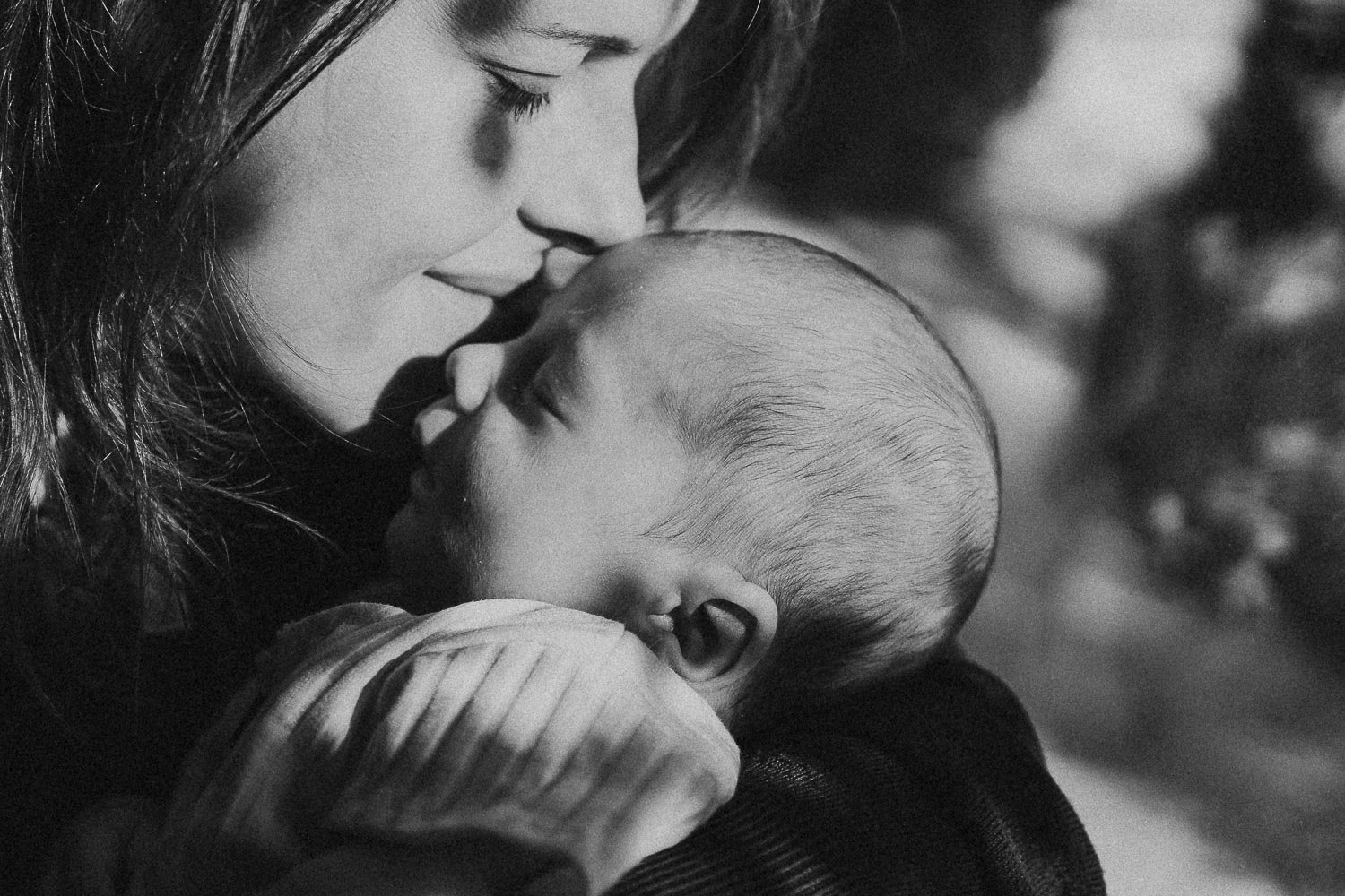 A mom smelling her baby's head, close to a window with some reflections of the tree leaves on her face during an in-home newborn photoshoot in Montreal