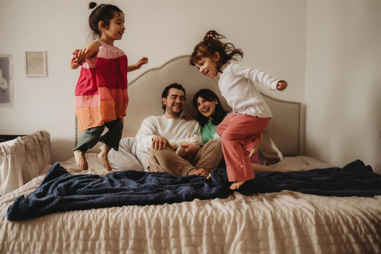 Girls jumping on the bed while parents are laughing and holding their newborn twin babies during an in-home newborn photoshoot in The West Island, Montreal