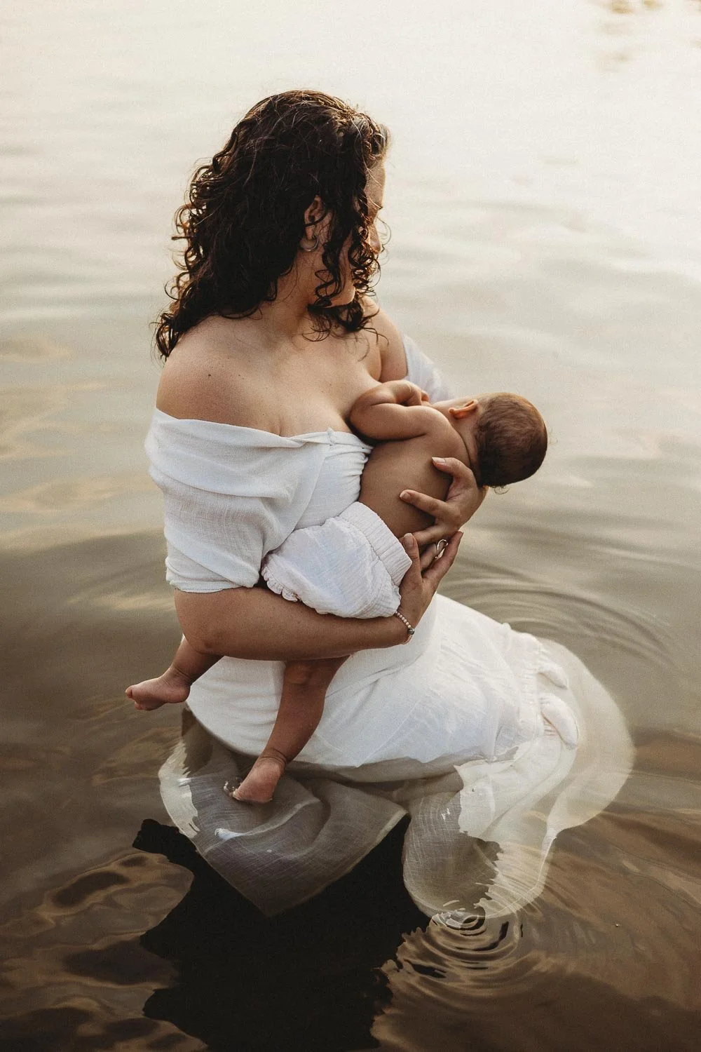 An outdoor motherhood photoshoot at Cap-Saint-Jacques portraying a mom in a white dress while breastfeeding her baby while sitting in the water, with a calm water background.