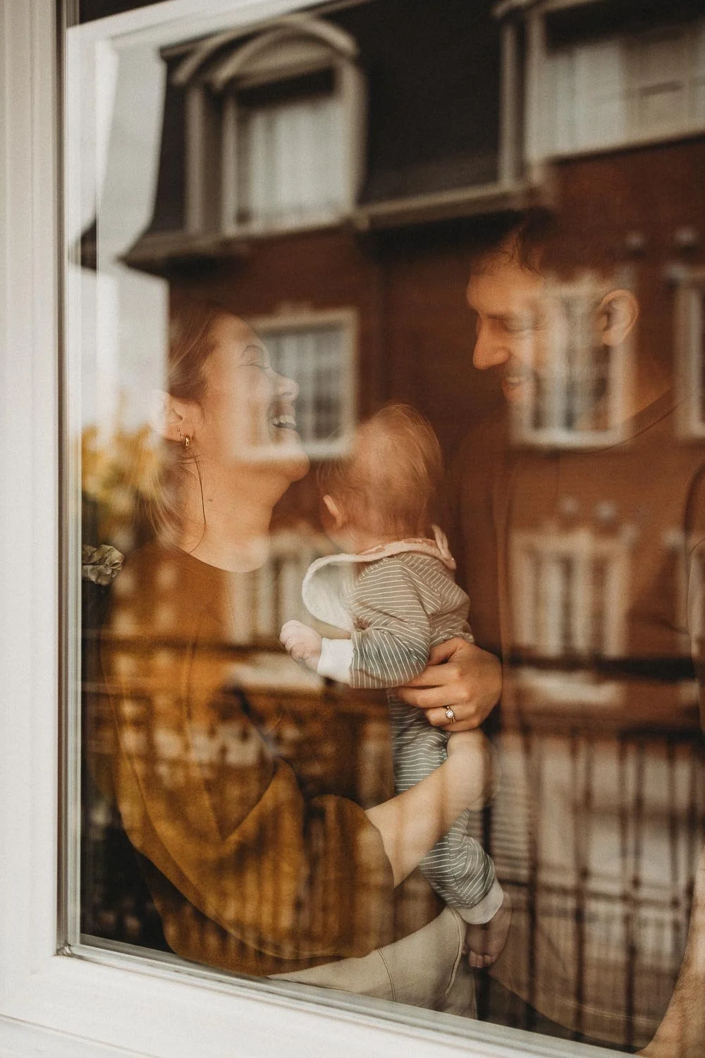 A woman and man looking at each other and smiling through a window, holding a baby between them, with reflections of bookshelves on the window glass captured during an in-home family photoshoot in West Montreal