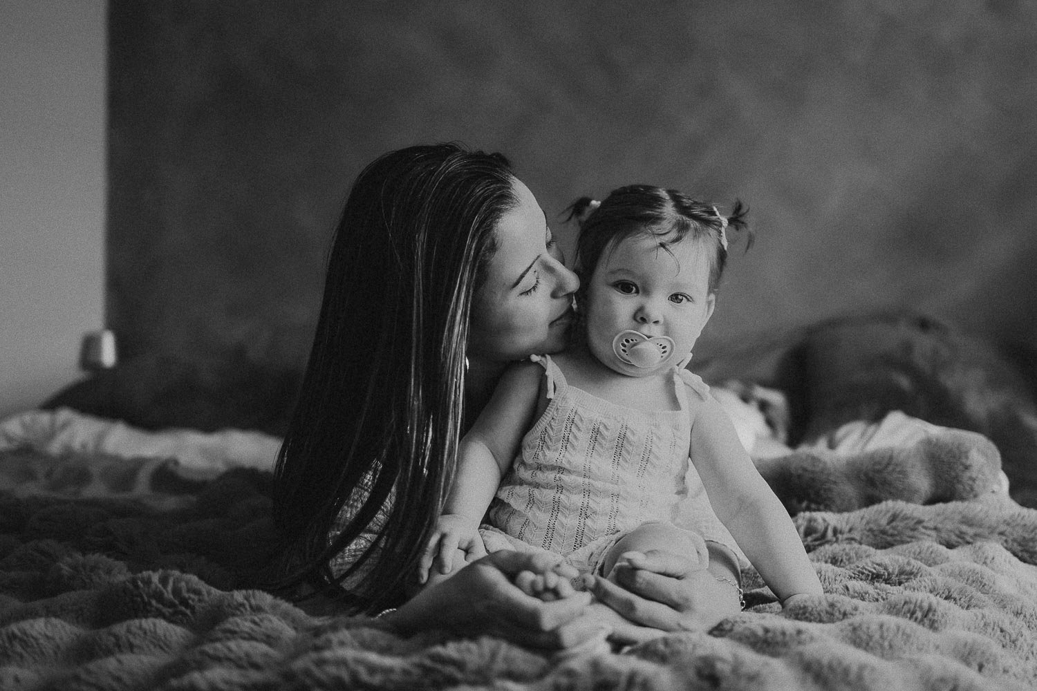 Mom laying down in bed next to her seated daughter. Mom is almost kissing her baby while the kid looks at camera. Captured at an in-home Motherhood Photoshoot in Montreal