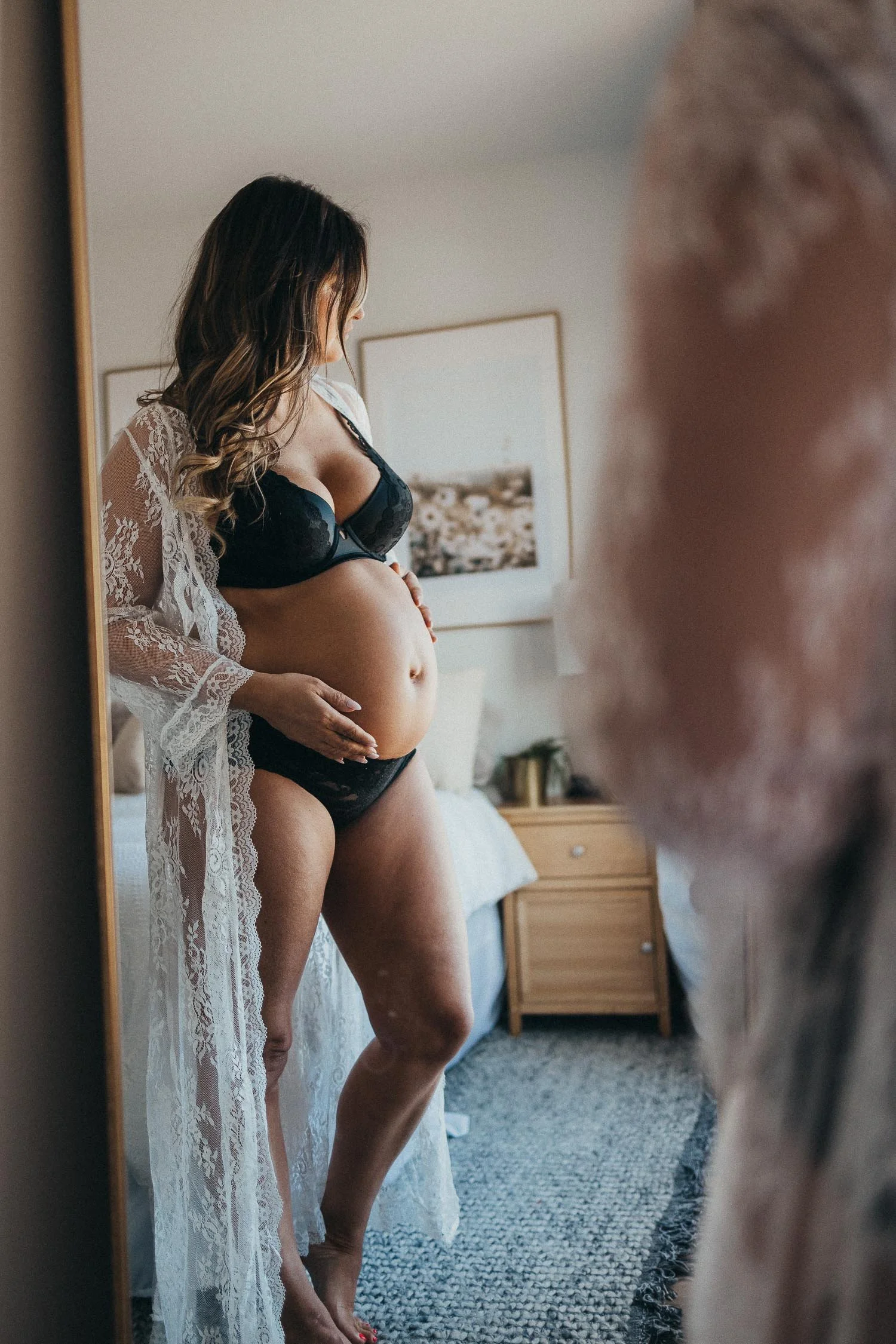 Pregnant woman in black lingerie and lace robe taking mirror selfie in bedroom, focused on her pregnant belly, with a blurred figure visible in the foreground.