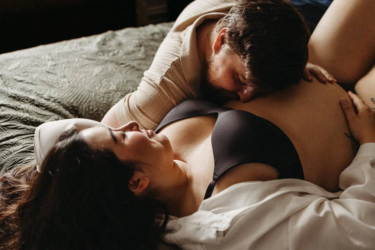 A couple lying on a bed, embracing and kissing, in a cozy, intimate in-home maternity photoshoot
