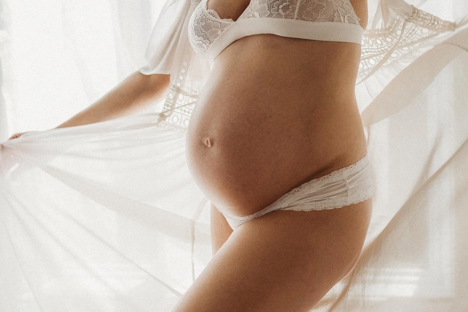 Close-up of a pregnant woman wearing white lace lingerie, holding a white lace umbrella, with soft natural lighting captured during an in-home boudoir maternity photoshoot in Montreal
