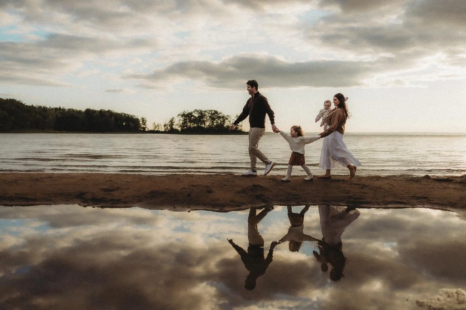 Family of four walking hand in hand along the shoreline at sunset, mirrored in the still water below captured during an outdoor photoshoot in Cap-Saint-Jacques Montreal