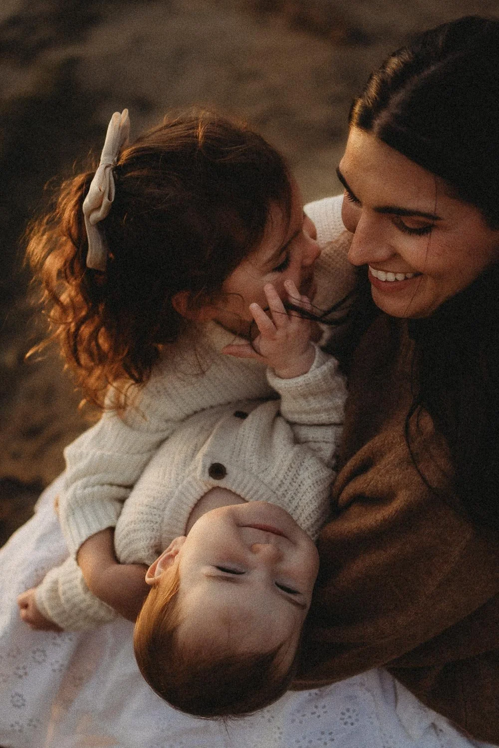 Toddler in a white bow kissing her mother's cheek while her baby sibling rests across her lap, autumn family session in Cap-Saint-Jacques Montreal