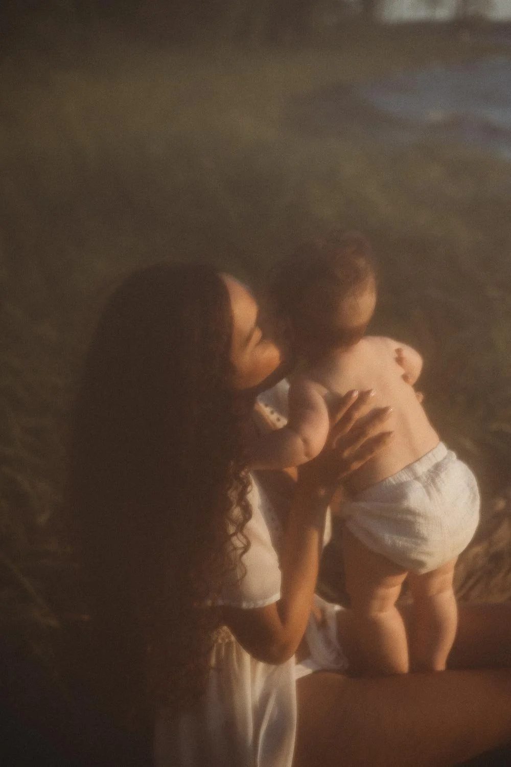 A woman with long curly hair is kissing a toddler on the cheek outside in a natural setting during a golden hour motherhood photo session at Cap-Saint-Jacques, Montreal