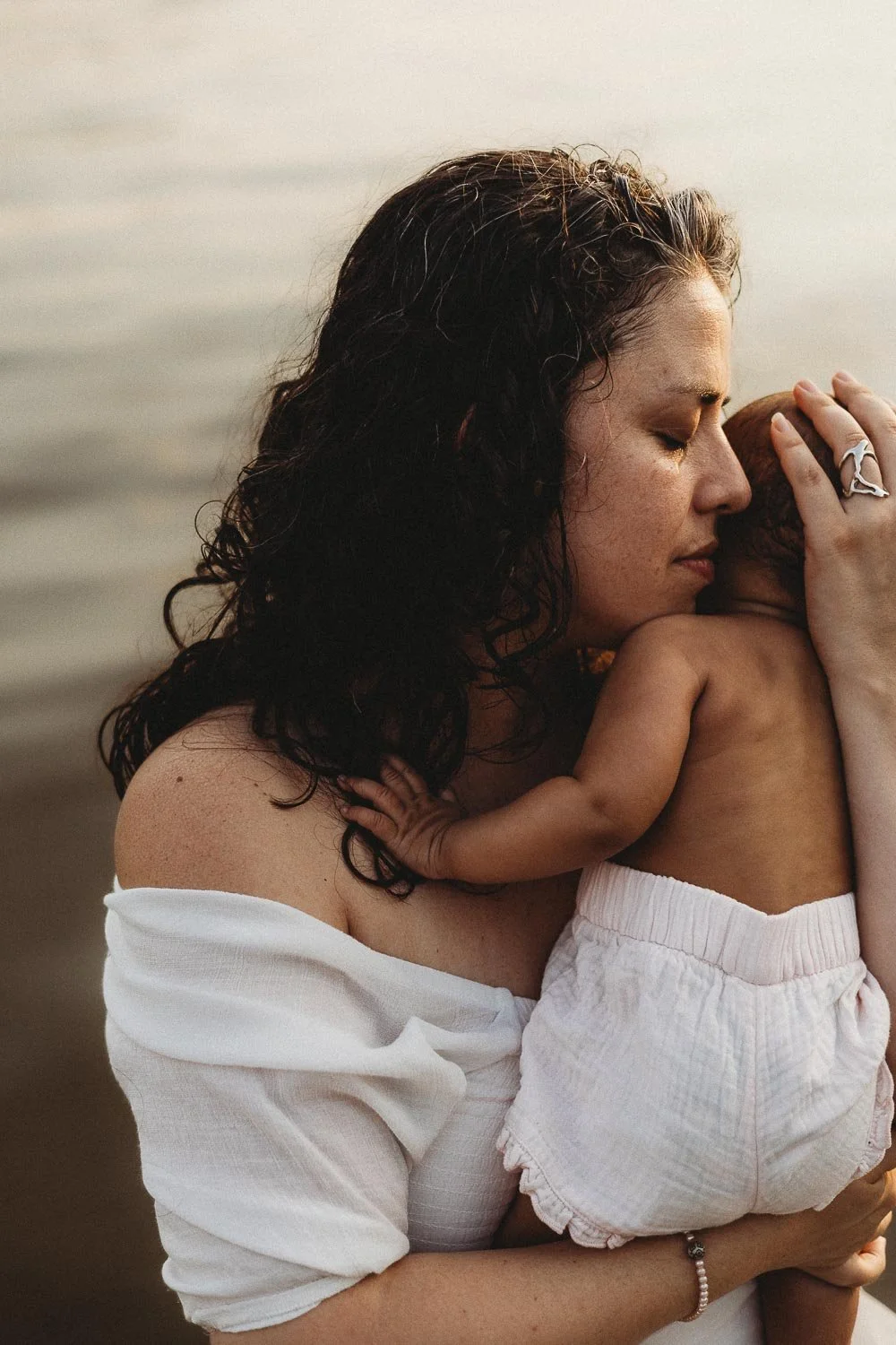 A woman with dark curly hair holding a small child close to her face at the beach.