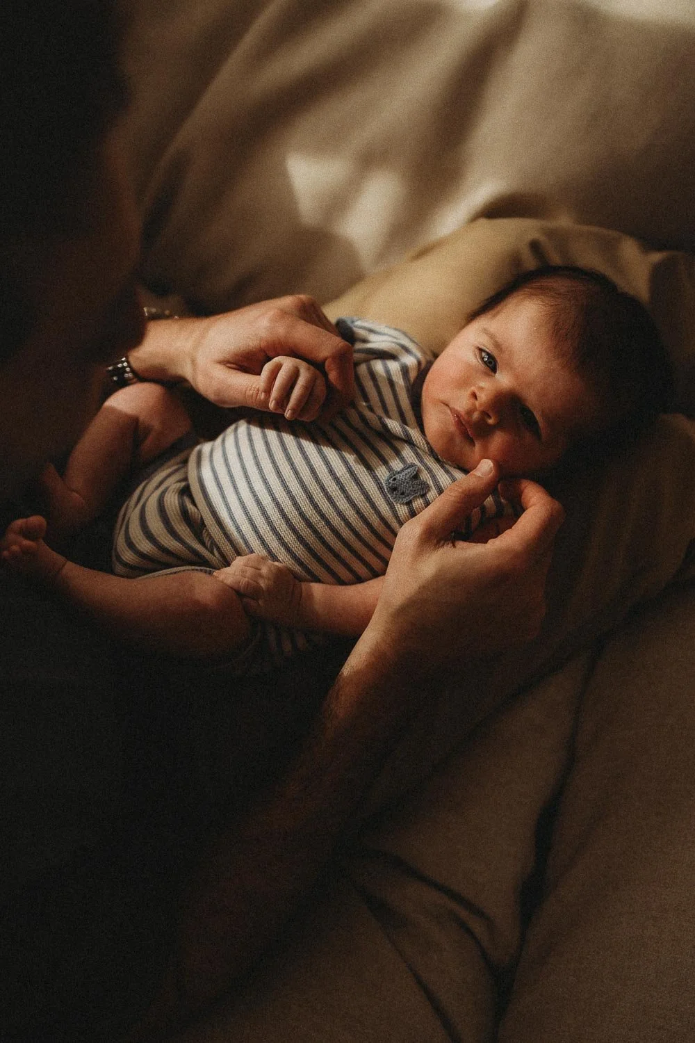 Baby boy laying down on his dad's lap while dad is holding one hand and caressing his little cheek in a mix of sunlight and shadows making it an intimate newborn photo, captured in Montreal