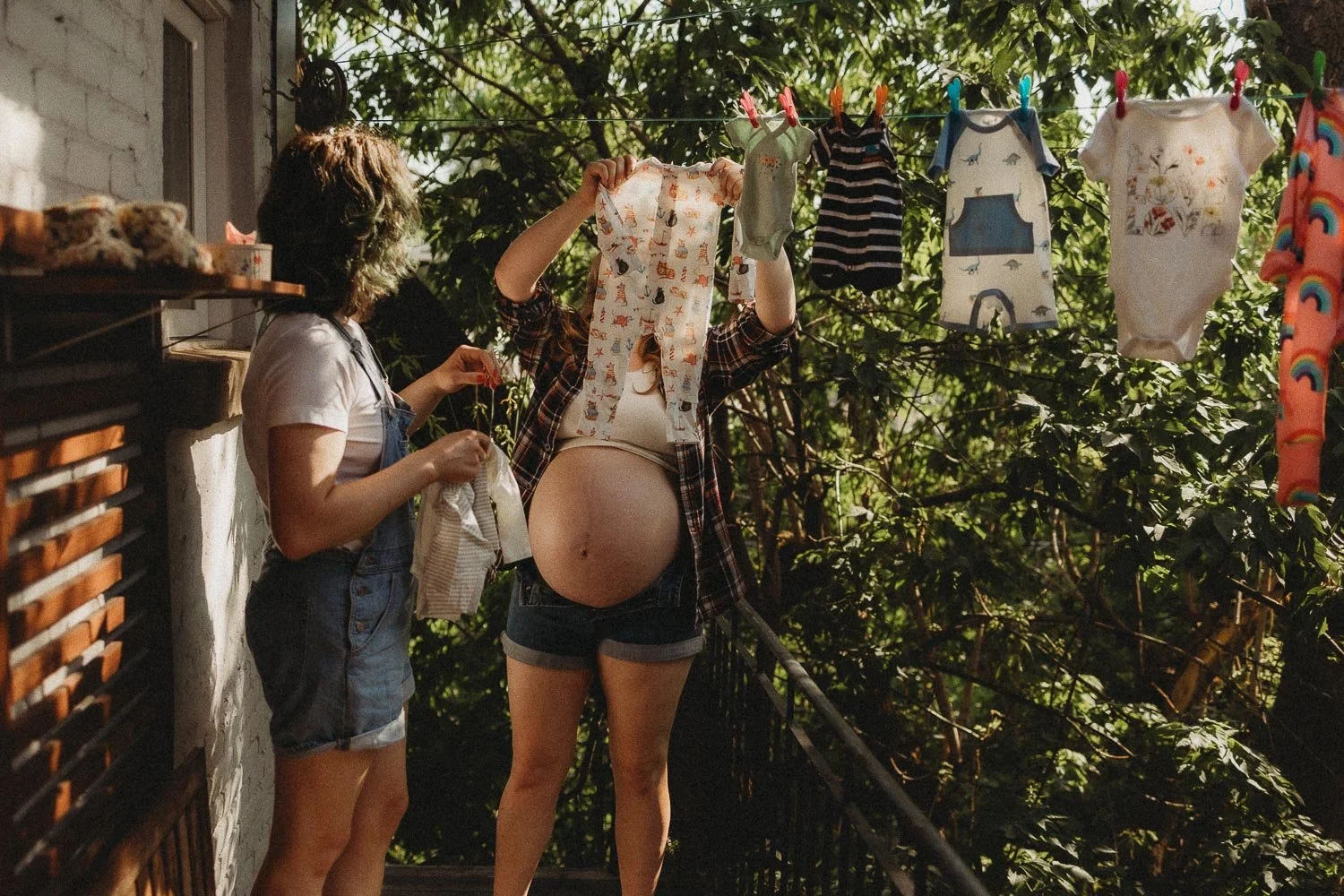 Pregnant mother pegging tiny baby clothes to the washing line with her partner outside their home before their baby arrives, captured during an in-home maternity photoshoot in Montreal