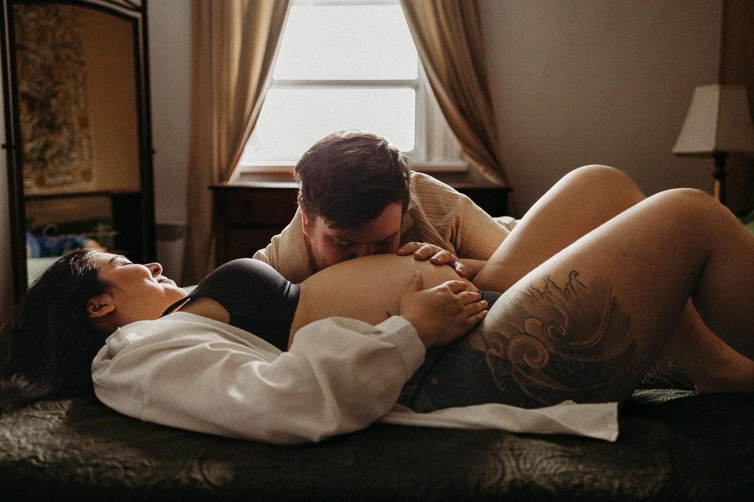 Pregnant woman laying down in bed while husband is gently kissing her belly and holding her at an in-home maternity photoshoot in La Salle, Montreal