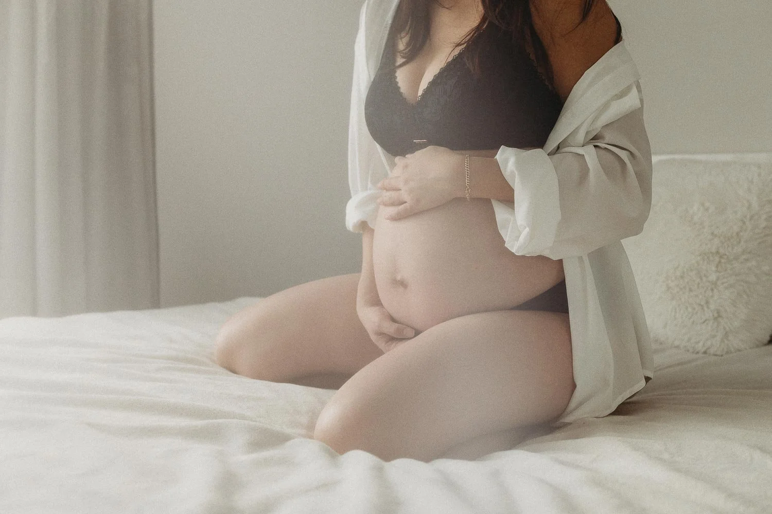 A person kneeling on a bed, wearing a black bra and white shirt, holding their pregnant belly with one hand, in a softly lit room.