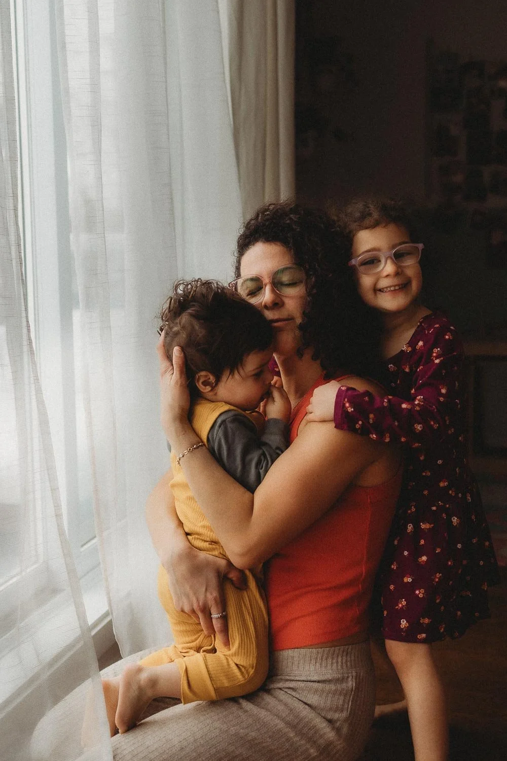 Jen Blanco hugging her two children, one boy and one girl, near a window with white curtains during a self-portrait motherhood session in Montreal.