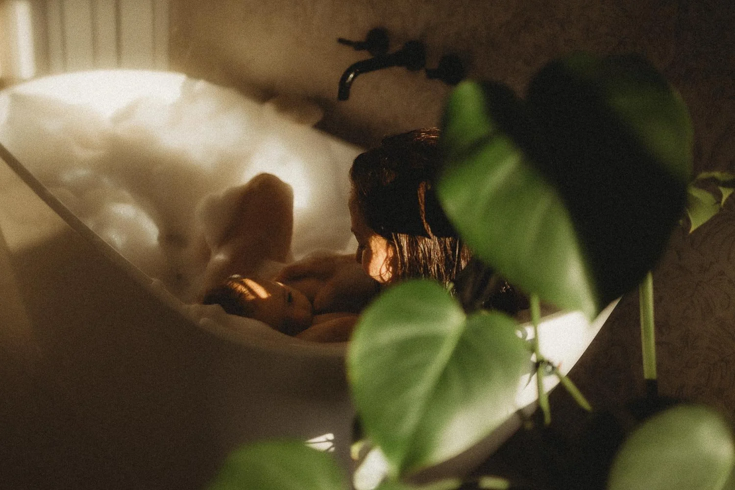 Person with wet hair lying in a white bathtub, partially obscured by green leaves of a plant in the foreground captured during an intimate motherhood photo session in Dorval, Montreal.