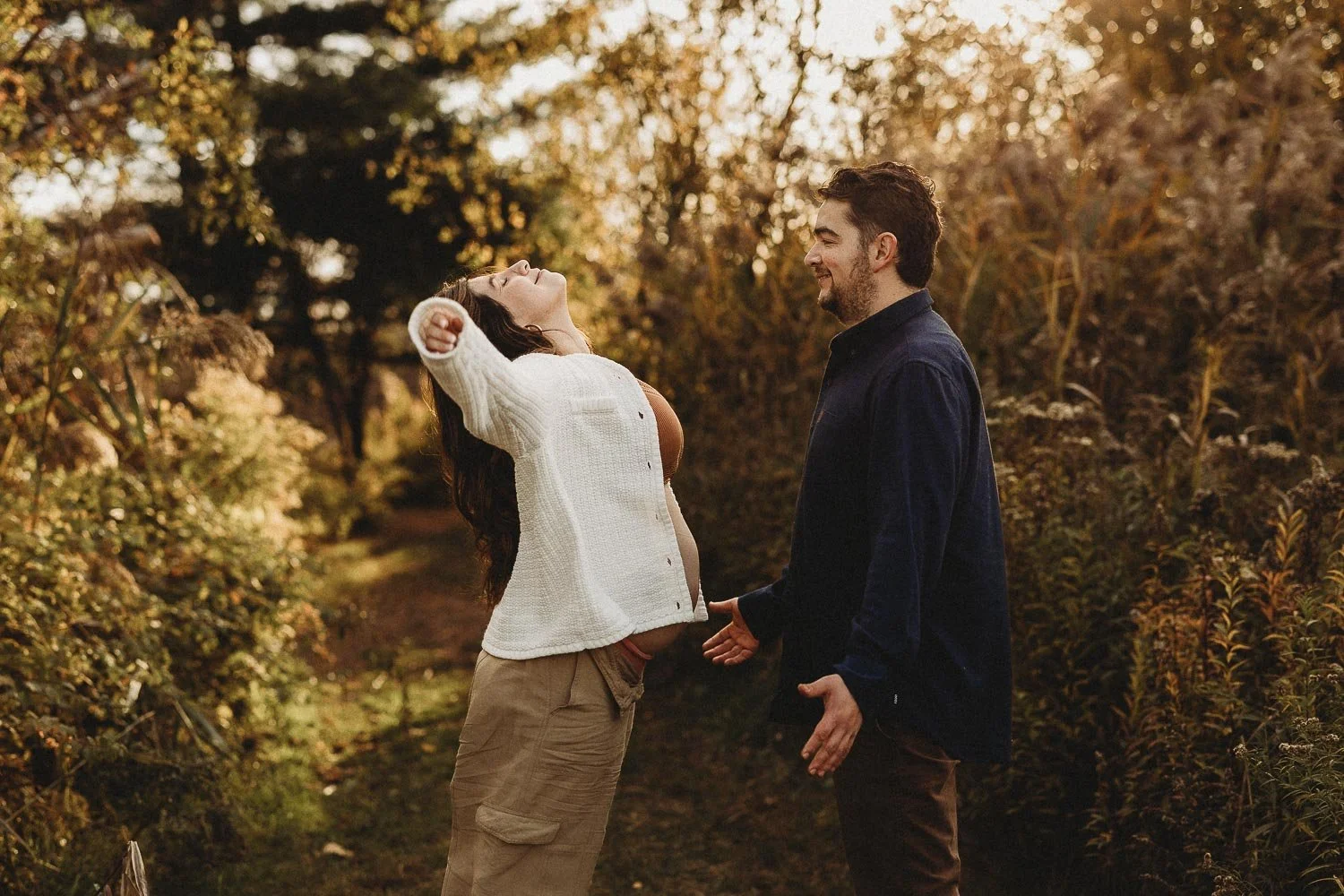 A man and woman standing on a trail surrounded by autumn-colored trees, smiling and holding hands, with the woman wearing a white cardigan and the man in a dark shirt during an outdoor maternity photoshoot in Boucherville Islands, near Montreal