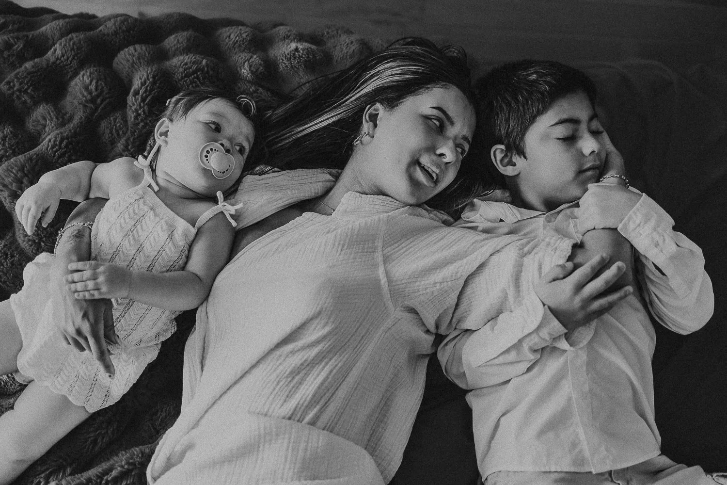 Mom and kids laying down on bed looking at each other and embracing in a hug, during a motherhood photo session in Montreal