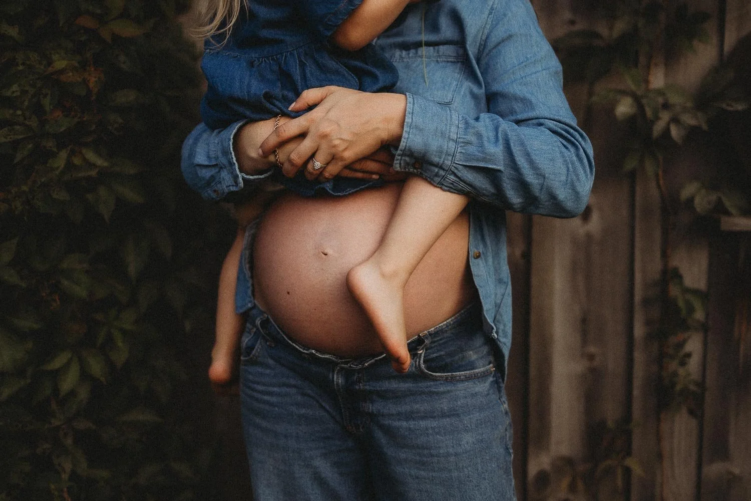 Close-up of a pregnant mama holding her toddler daughter, we can see the belly and the legs and feet of the girl, captured during an outdoor maternity photoshoot at Le Verger Du Flanc Nord in Mont Saint-Hilaire
