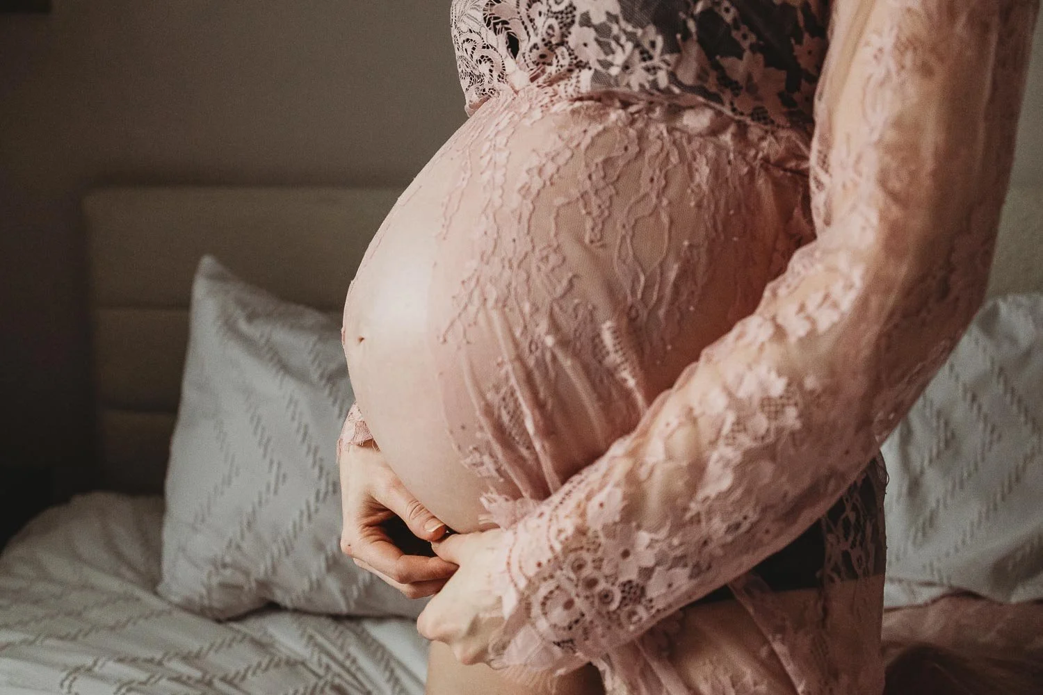 A closed-up photo of a pregnant woman's belly, sh's gently holding her hands and wearing a lace robe that reveals her belly, captured during an in-home photoshoot in West Island Montreal