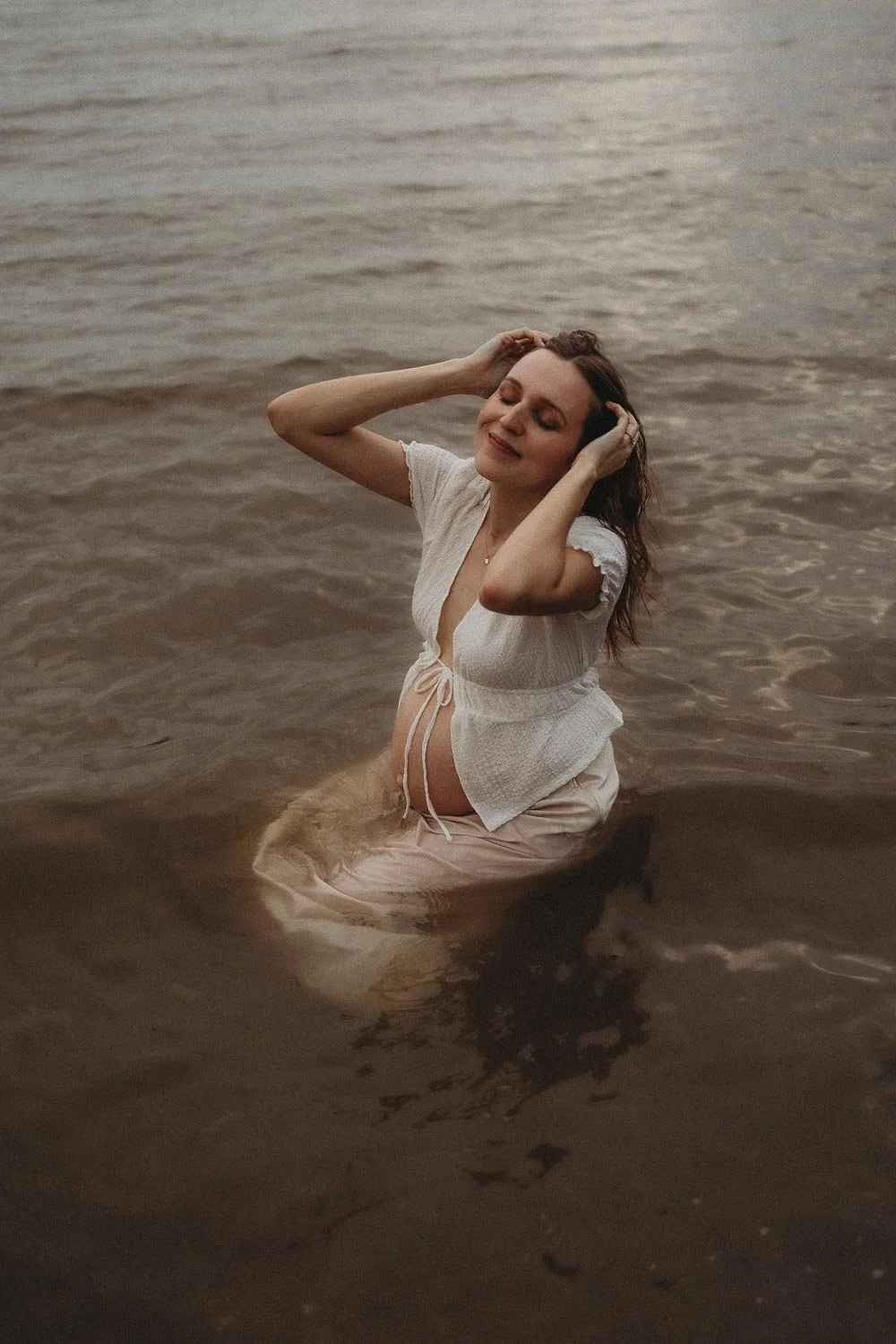Pregnant woman seated in the water while wearing maternity clothes and holding her hair with a smirk in her face, captured during an outdoor maternity photoshoot in Cap-Saint-Jacques, Montreal