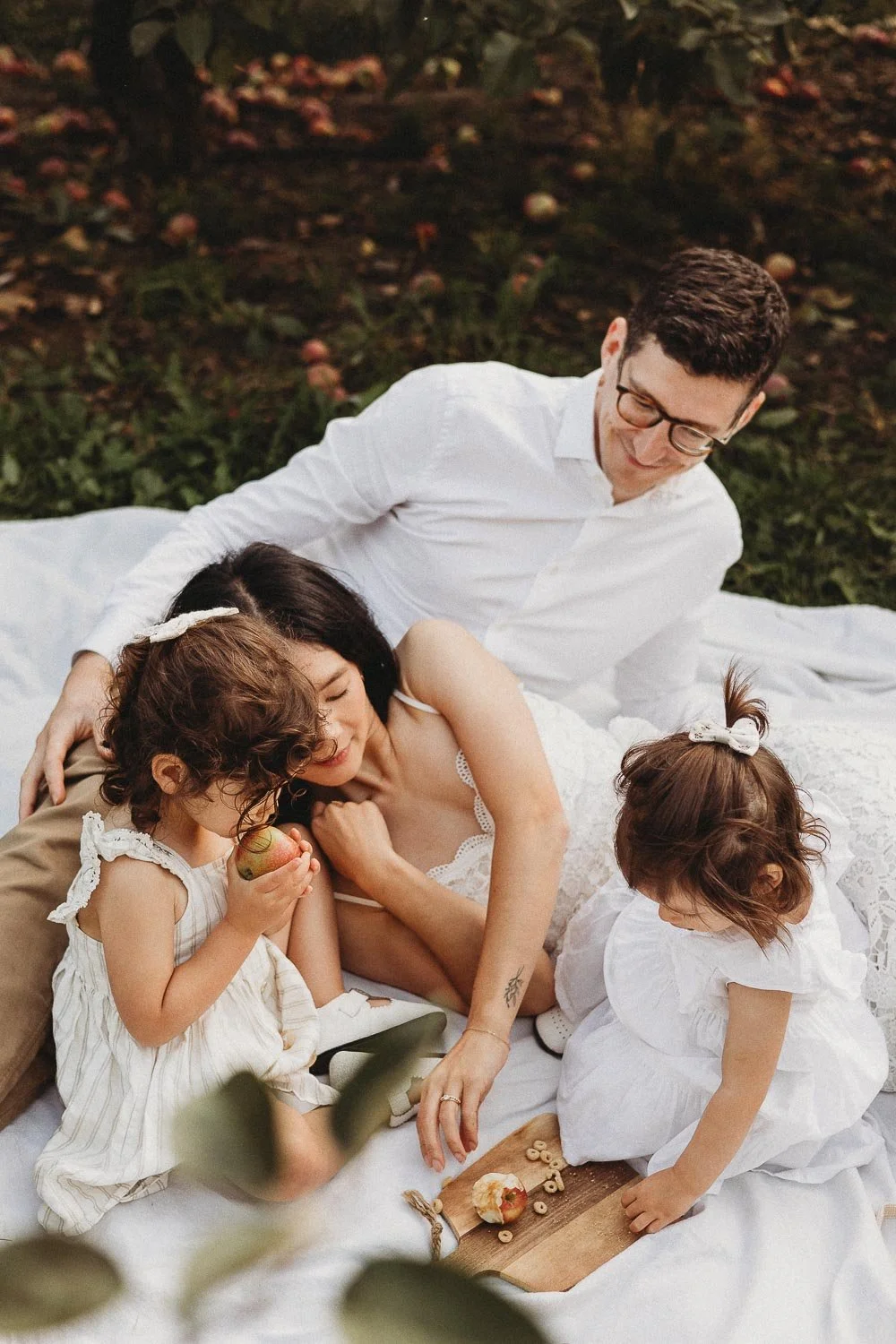 A family of four having a picnic outdoors on a blanket, including a man, woman, and two young girls, with trees and fallen apples in the background during an outdoor family photoshoot at the Verger du Flanc Nord