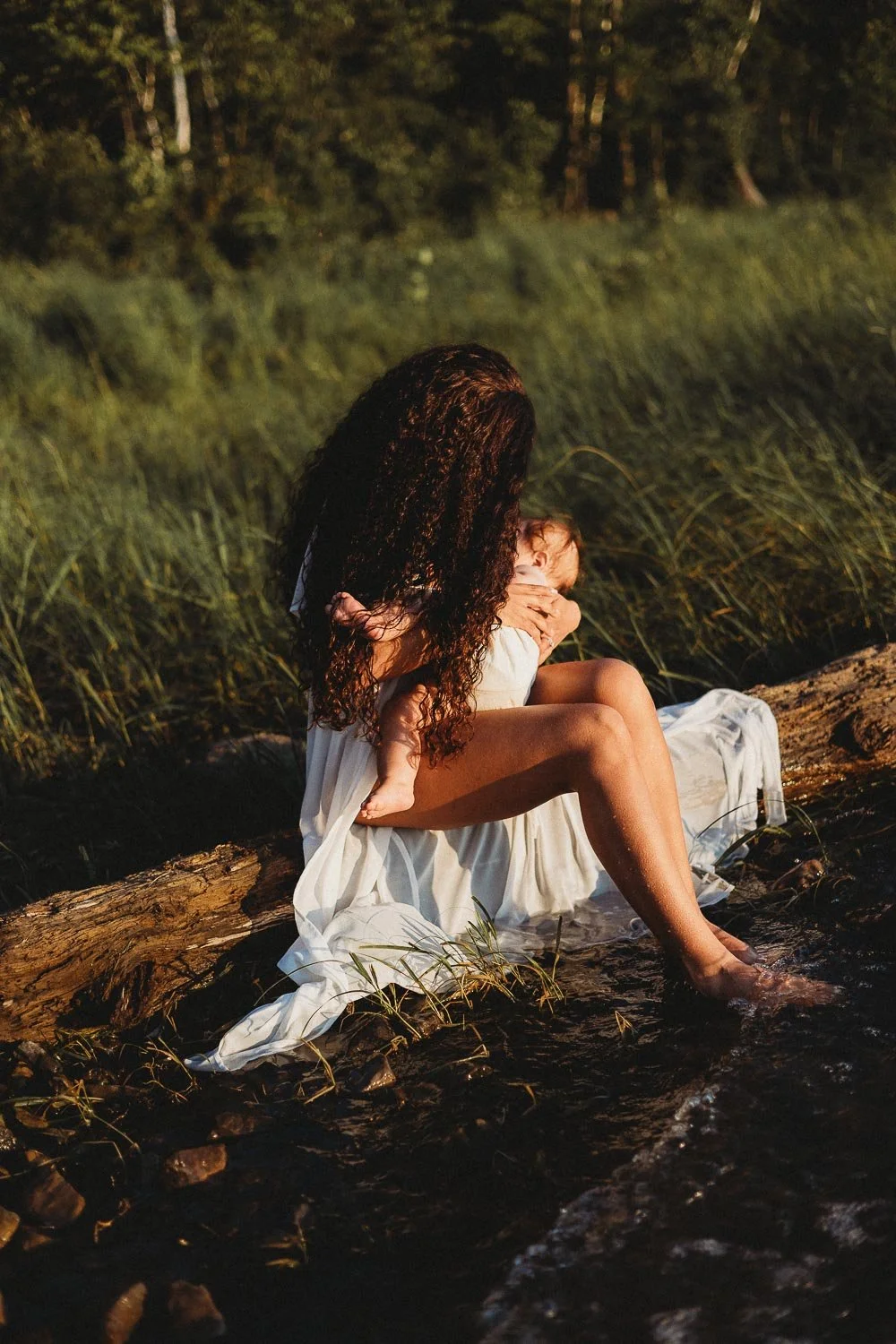 A woman with long curly hair sitting on a log in a river, holding a young child in her arms, surrounded by green foliage at sunset.