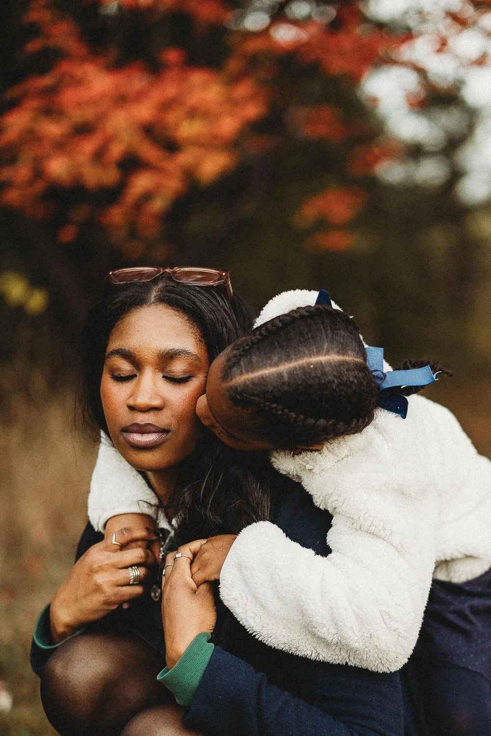 An outdoor motherhood photoshoot in Maisonneuve Park portraying a woman with glasses on her head and rings on her fingers is hugging a young girl with braided hair decorated with a blue ribbon outdoors with blurred autumn leaves in the background.