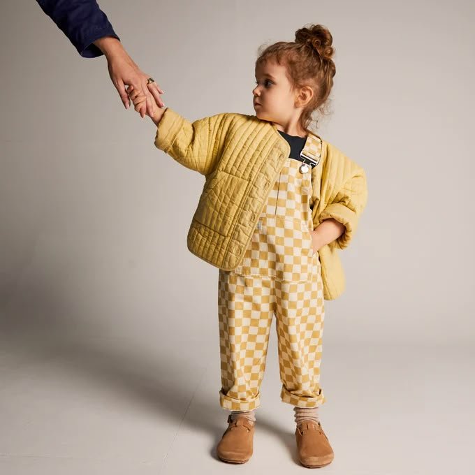 Young child in yellow quilted jacket and checkered overalls holds an adult's hand.
