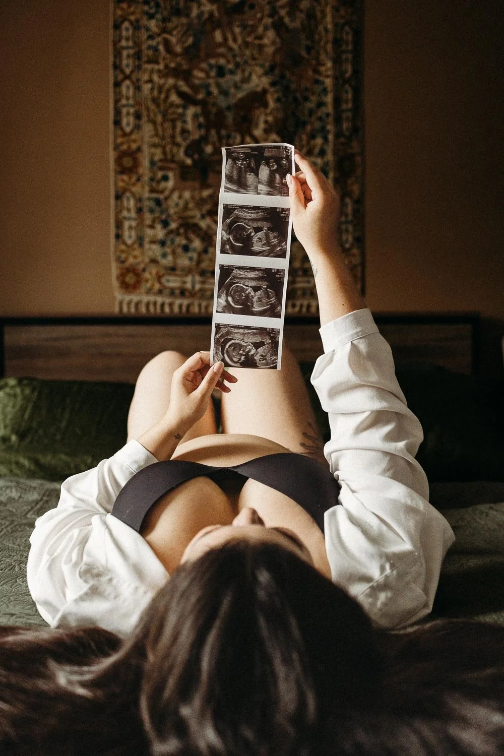 A woman lying on a bed holding ultrasound images of pregnancy, with a tapestry hanging on the wall in the background.