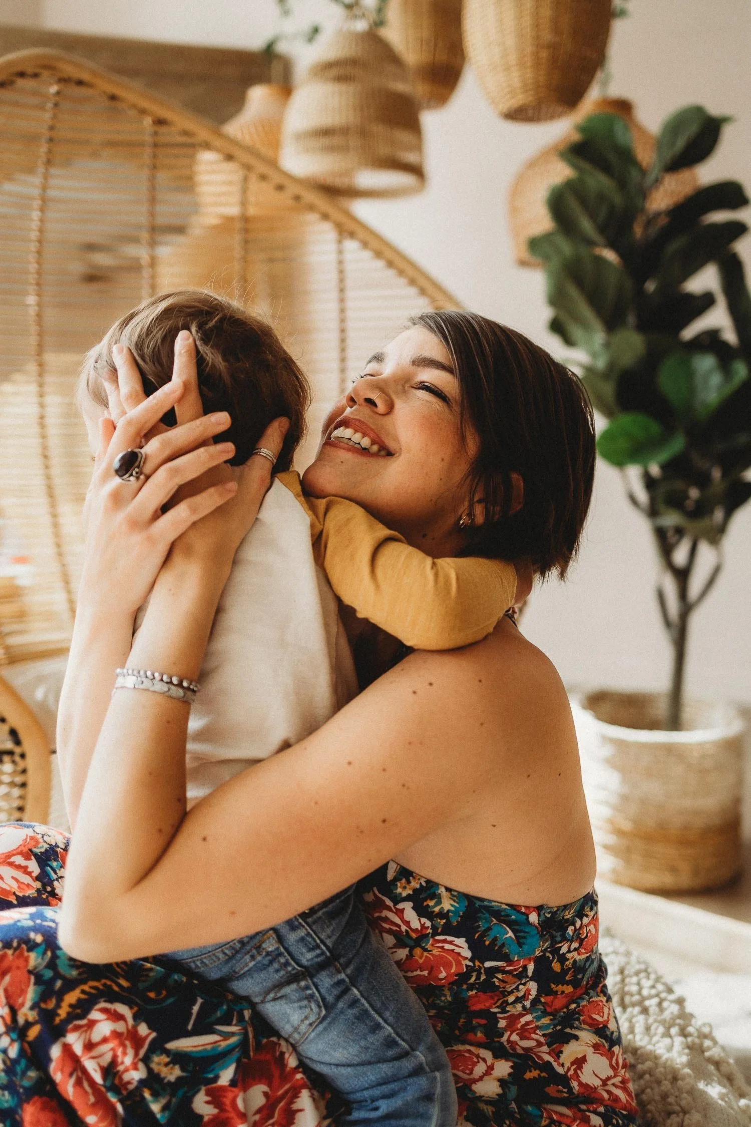 A woman happily hugs a young child indoors, with a decorative wicker background and a potted plant visible during a motherhood photoshoot in Montreal.