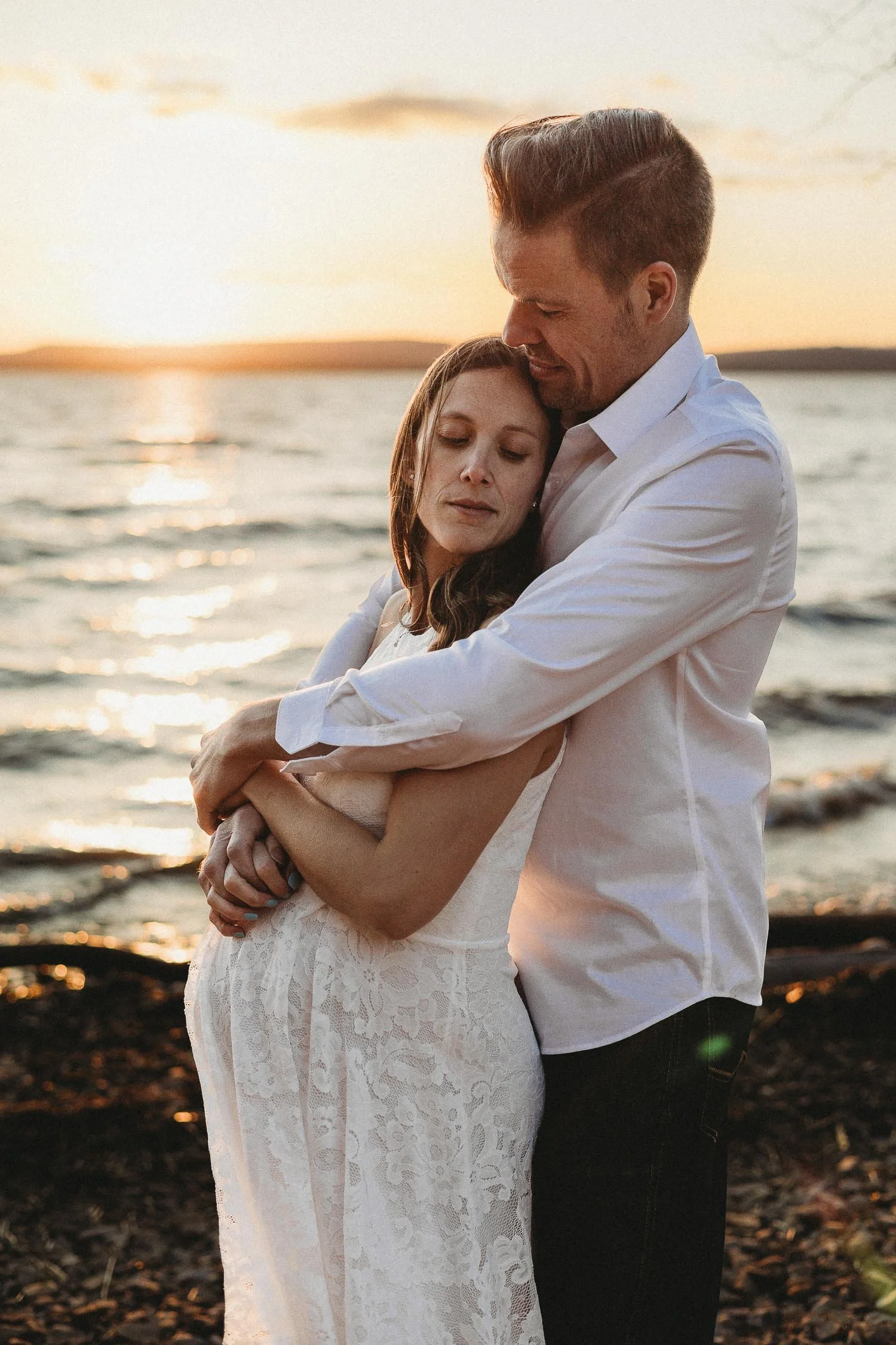 A couple embracing by the water at sunset, with the woman in a white lace dress and the man in a white shirt and dark pants.