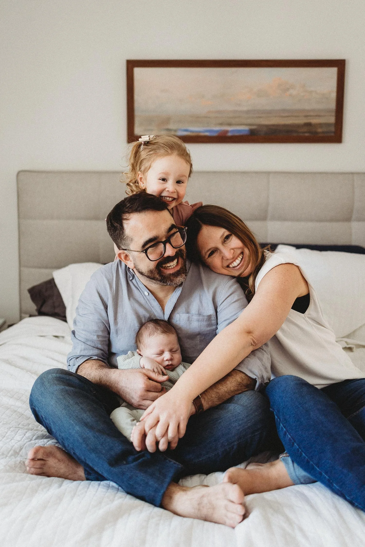 Family of four sitting on a bed, smiling, with a toddler on the man's shoulders and a newborn in the man's arms captured during an in-home newborn photoshoot in TMR, Montreal.