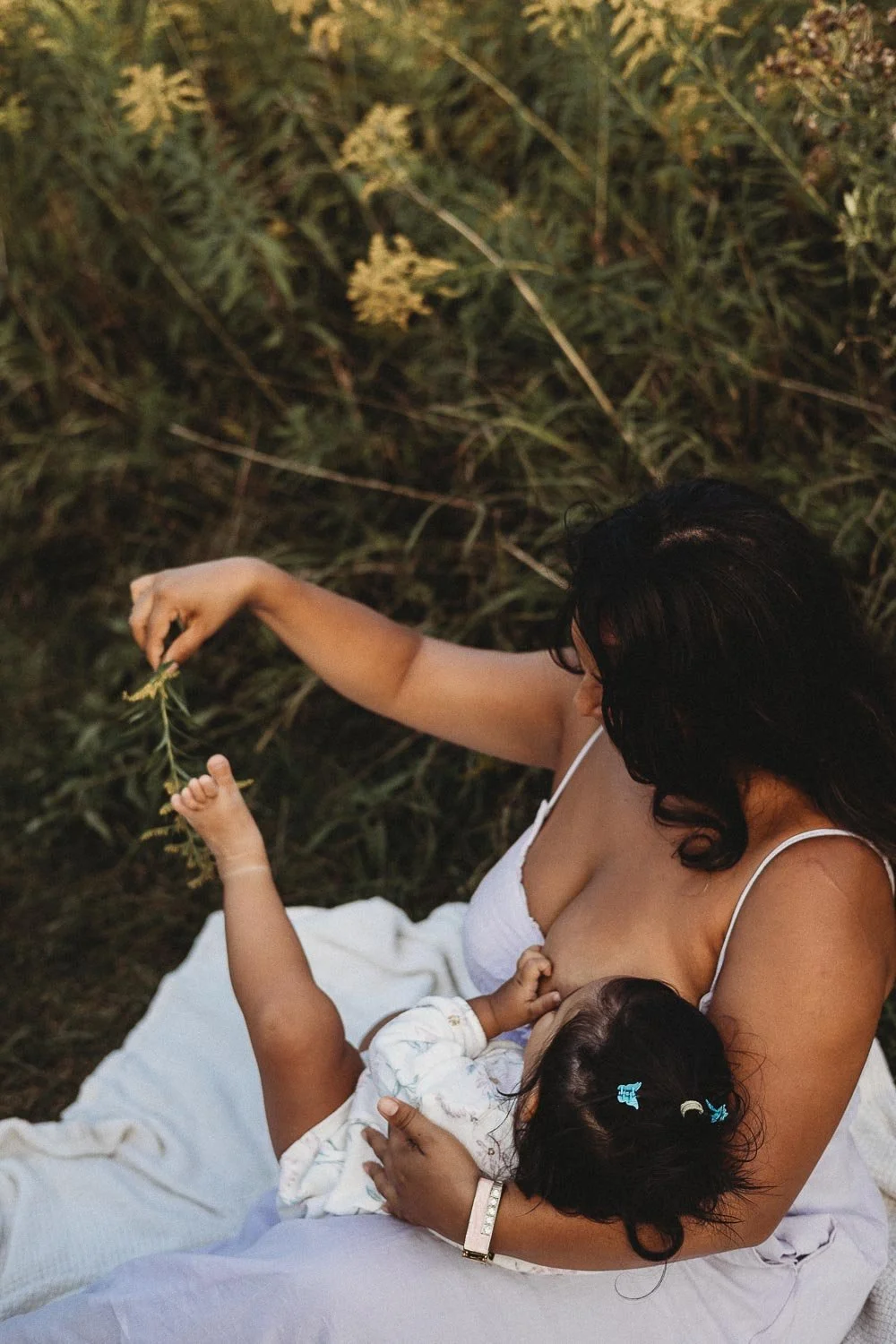 A woman is sitting outdoors on a white blanket, breastfeeding a baby. The woman is reaching out with her hand to hold a small plant or flower. The background features tall grass or plants.