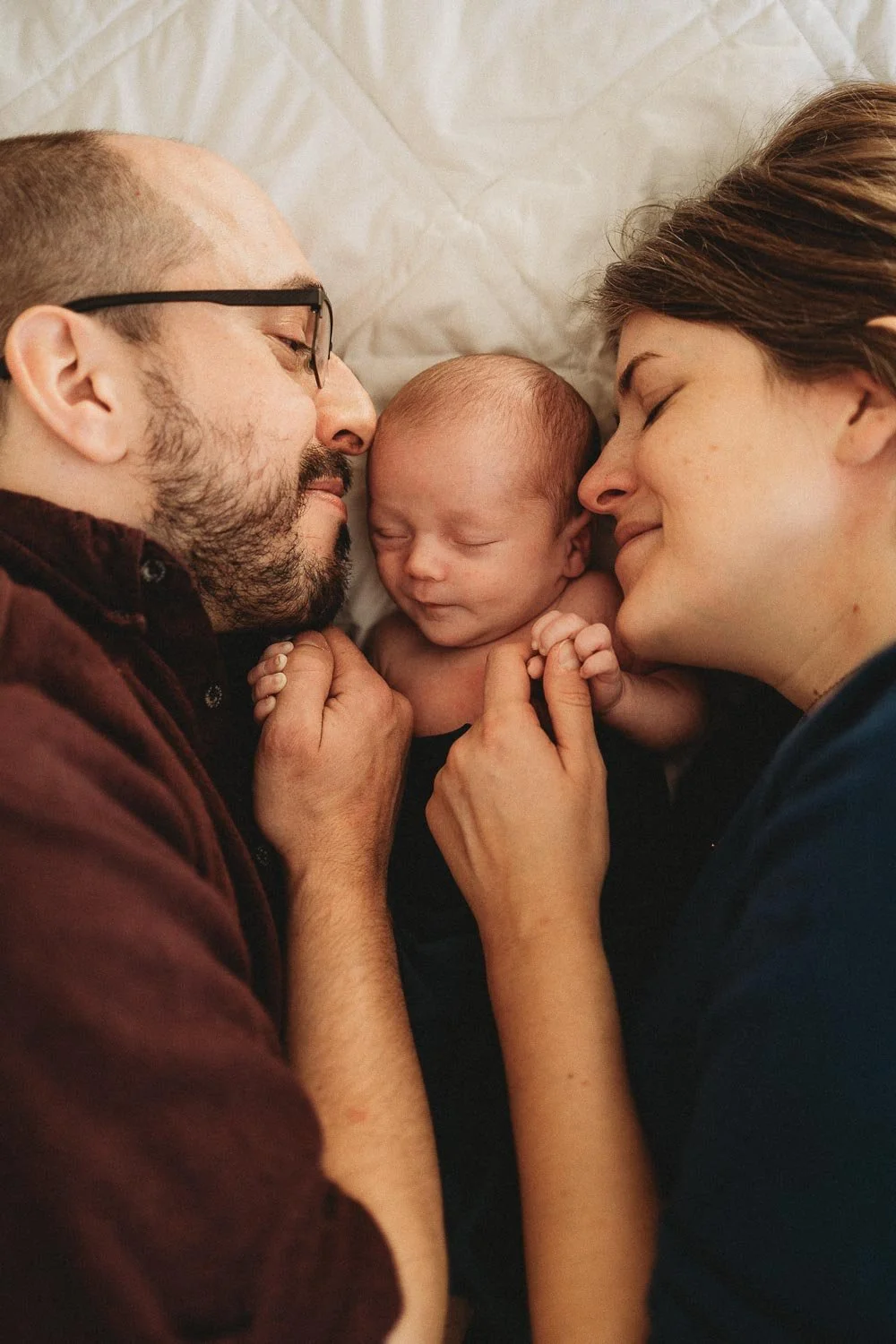 A family of three, a man, a woman, and a newborn baby, lying in bed close together and the parents are holding their baby's hands during an in home newborn photoshoot in Montreal