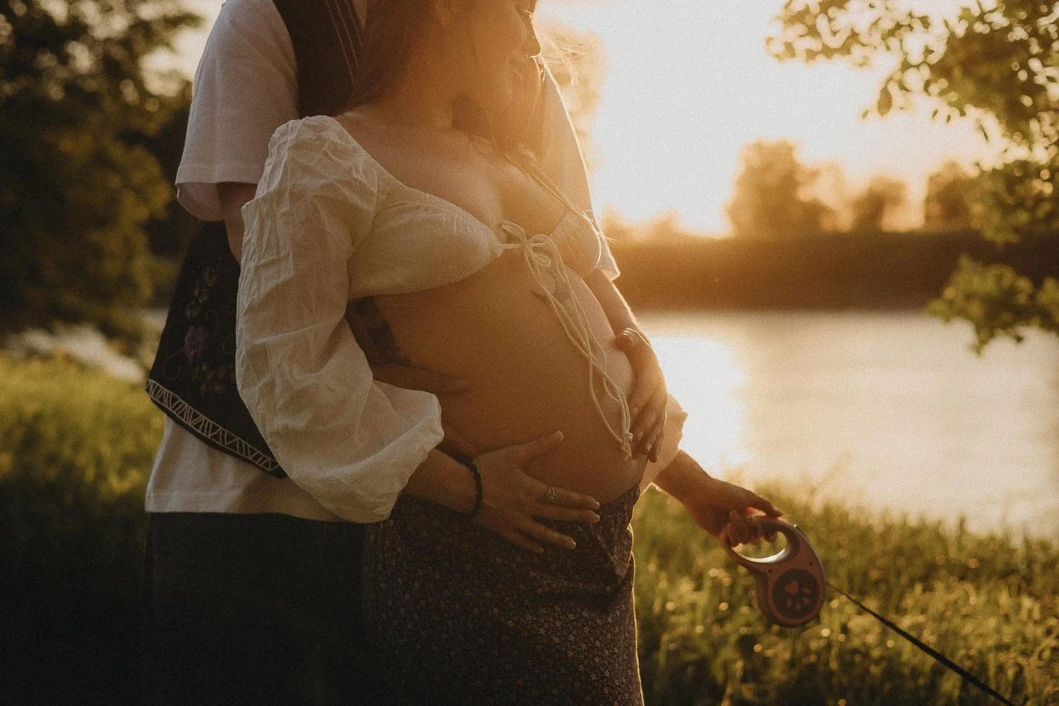 A romantic photo of a pregnant woman being held from the back by her partner, both holding her pregnant belly with the golden hour sun shinning on them at an outdoor maternity photo session in Boucherville Island, Quebec